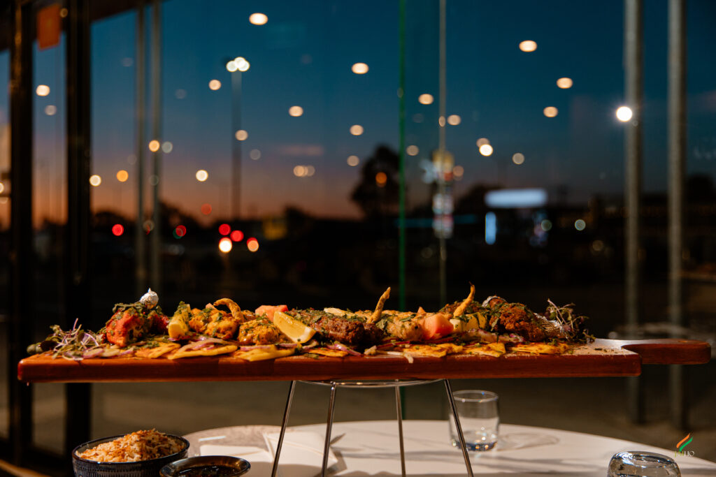 Premium mixed grill platter served on a wooden board with rice and appetizers in a restaurant during sunset, featuring warm ambient lighting and bokeh background.